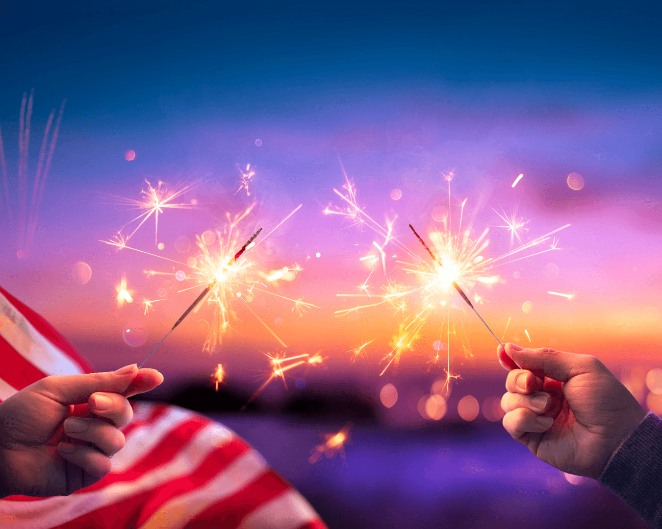 Two hands holding sparklers together with lake, American flag, and fireworks in background during sunset.