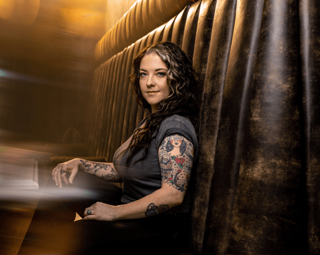 Ashley McBryde sitting in bar booth, looking to her left, smiling at cmera.